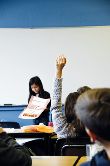 Klassrumsbild - en flikca visar upp ett arbete och en elev räcker upp handen. 